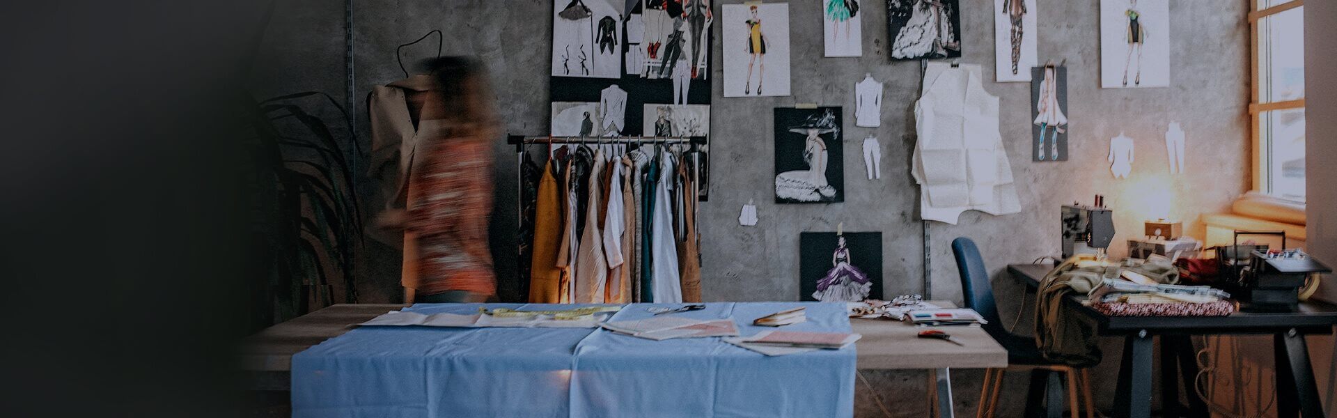 Image of a fashion design studio, with drawings on the wall, a desk with a sewing machine, and work top for cutting fabric